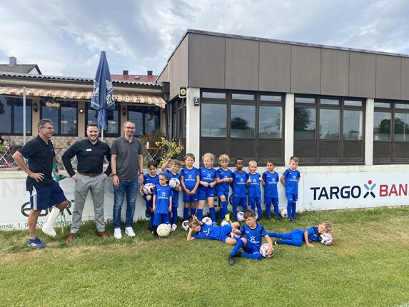 Eine Gruppe von Kindern in blauen Fußballtrikots steht vor einem Gebäude. Zwei Erwachsene, einer in schwarzem Hemd und einer in grauem Hemd, stehen neben den Kindern. Die Kinder halten Fußball und posieren. Im Hintergrund ist ein Schild mit "TARGO BANK" zu sehen. Es ist ein bewölkter Tag.