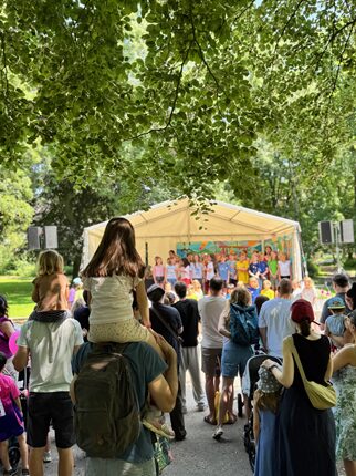Eine Menschenmenge versammelt sich im Park, um eine Aufführung auf einer Bühne zu sehen. Auf der Bühne stehen mehrere Darsteller, die in bunten Kostümen gekleidet sind. Im Vordergrund sitzt ein Kind auf den Schultern eines Erwachsenen, während andere Zuschauer aufmerksam zuschauen. Die Szene ist von Bäumen umgeben, die Schatten spenden.