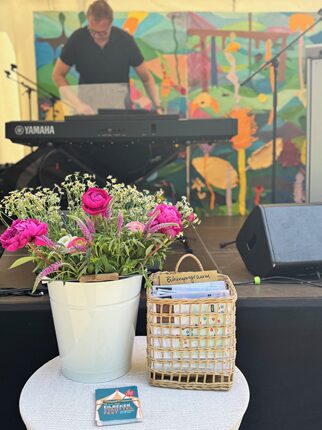 Ein Musiker spielt an einem Yamaha-Piano vor einem bunten Hintergrund mit Tieren und Pflanzen. Im Vordergrund steht ein Tisch mit einem weißen Blumentopf, der rosa Rosen und kleine weiße Blumen enthält, sowie einem geflochtenen Korb mit Notizen und einer Karte.