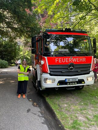 Ein Feuerwehrmann steht neben einem roten Feuerwehrfahrzeug auf einem gepflasterten Weg in einem Park. Das Fahrzeug hat die Aufschrift 
