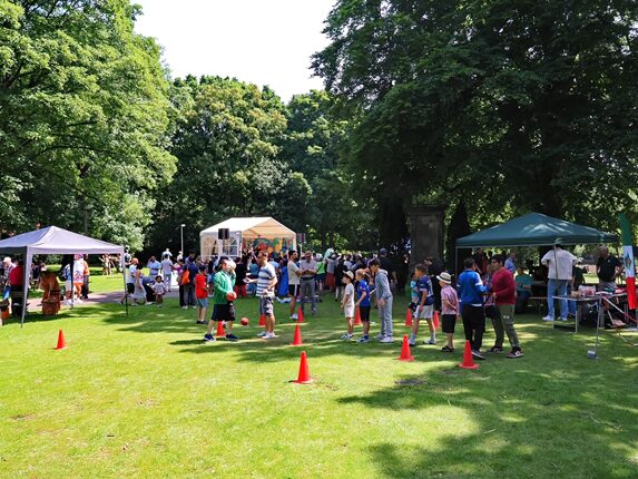 Eine belebte Parkveranstaltung mit vielen Menschen, darunter Kinder und Erwachsene. Zelte in verschiedenen Farben stehen auf dem Rasen, und es gibt orangefarbene Pylonen auf dem Boden. Im Hintergrund sieht man eine Bühne mit einer großen Menschenmenge. Bäume umgeben den Bereich und es ist sonnig.