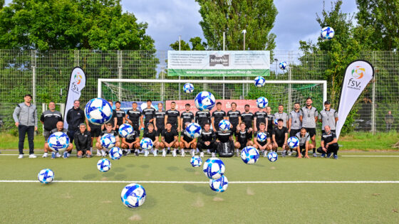 Eine Gruppe von Fußballspielern und Trainern posiert auf einem Sportplatz, umgeben von mehreren schwebenden Fußbällen. Sie tragen schwarze Trikots und stehen in einer Reihe vor einem Fußballtor. Im Hintergrund sind Banner und Bäume sichtbar. Es gibt zwei Fahnen mit dem Logo 'fsv Waiblingen'.