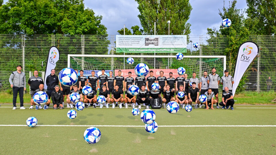 Eine Gruppe von Fußballspielern und Trainern posiert auf einem Sportplatz, umgeben von mehreren schwebenden Fußbällen. Sie tragen schwarze Trikots und stehen in einer Reihe vor einem Fußballtor. Im Hintergrund sind Banner und Bäume sichtbar. Es gibt zwei Fahnen mit dem Logo 'fsv Waiblingen'.