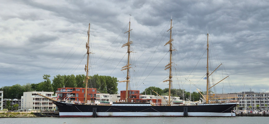 Ein großes Segelschiff mit mehreren Masten liegt im Hafen. Es hat einen schwarzen Rumpf und goldene Masten. Im Hintergrund sind moderne Wohngebäude zu sehen, während der Himmel bewölkt ist.