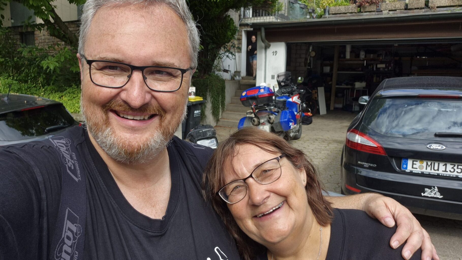 Ein Mann mit Brille und grauem Bart lächelt in die Kamera, während er eine Frau mit Brille umarmt. Im Hintergrund sind Motorräder und Autos sichtbar, darunter ein schwarzes Auto und ein blauer Roller. Das Bild scheint im Freien vor einem Wohnhaus aufgenommen worden zu sein.