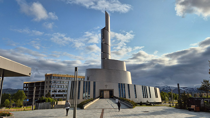 Eine moderne Kirche mit einer hohen, schmalen Spitze und einer runden Basis. Der Eingang ist zentral platziert, umgeben von großen Fenstern. Im Hintergrund ist ein mehrstöckiges Gebäude zu sehen. Die Umgebung ist grün, mit Bäumen und gepflasterten Wegen unter einem teilweise bewölkten Himmel.