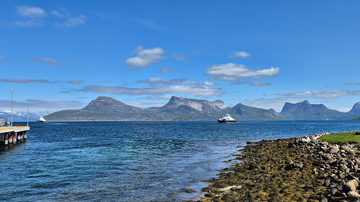 Ein klarer Himmel mit wenigen Wolken über einem ruhigen Gewässer, das von Bergen umgeben ist. Ein Boot fährt durch das Wasser, während im Vordergrund ein Steg und ein Kiesstrand sichtbar sind. Die Landschaft ist grün und felsig, mit klarem, blauem Wasser, das die Bergkulisse reflektiert.
