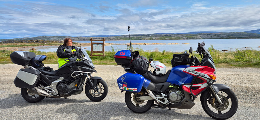 Zwei Motorräder stehen an einem ruhigen Ufer. Ein schwarzes Motorrad mit Koffer und ein blau-rotes Motorrad mit Gepäck sind sichtbar. Eine Person in gelber Kleidung steht neben dem schwarzen Motorrad und schaut in die Ferne. Im Hintergrund sind sanfte Hügel und ein ruhiger See mit Wolken am Himmel zu sehen.