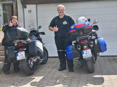 Zwei Personen stehen neben zwei Motorrädern in einer Einfahrt. Eine Frau gibt ein Daumen-hoch-Signal, während ein Mann lächelt und ebenfalls ein Daumen-hoch-Signal zeigt. Hinter ihnen ist eine Garagentür sichtbar. Die Motorräder haben jeweils Gepäcktaschen und sind in verschiedenen Farben lackiert.