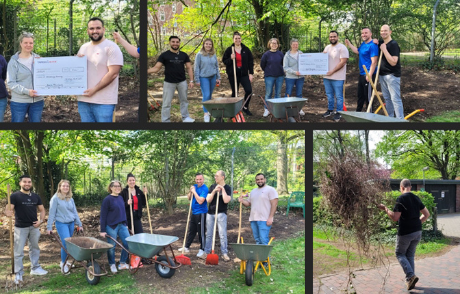 Eine Gruppe von Menschen steht in einem Garten mit Schaufeln und Schubkarren. Einige halten ein großes Scheck-Schild, während andere lächeln und posieren. Bäume und Sträucher sind im Hintergrund sichtbar, und die Gruppe scheint an einem Gemeinschaftsprojekt zu arbeiten.