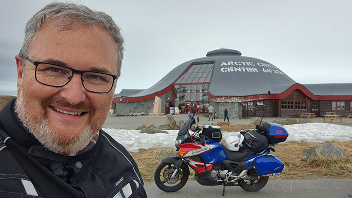 Ein Mann mit Brille und grauem Bart lächelt vor dem Arctic Center. Im Hintergrund sieht man das moderne Gebäude mit einem geschwungenen Dach und großen Fenstern. Vor dem Gebäude steht ein Motorrad mit Gepäck. Es gibt Schnee und Gras im Vordergrund.