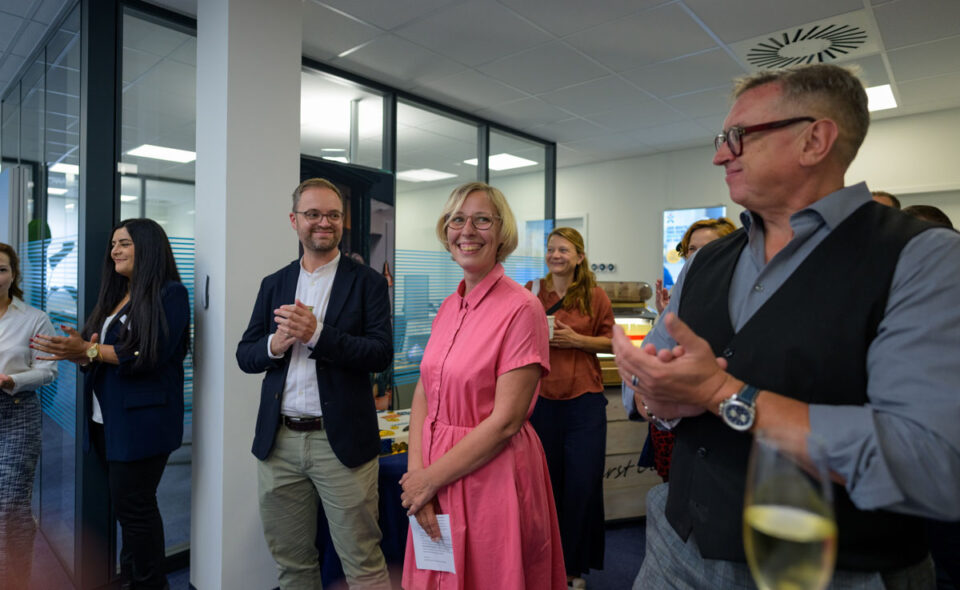 Eine Gruppe von Menschen applaudiert in einem modernen Büro. In der Mitte steht eine Frau mit blonden, kurzen Haaren und einer rosa Bluse, die lächelt. Neben ihr steht ein Mann mit Brille in einem dunklen Anzug. Weitere Personen sind im Hintergrund zu sehen, einige halten Gläser. Die Atmosphäre ist feierlich und freundlich.