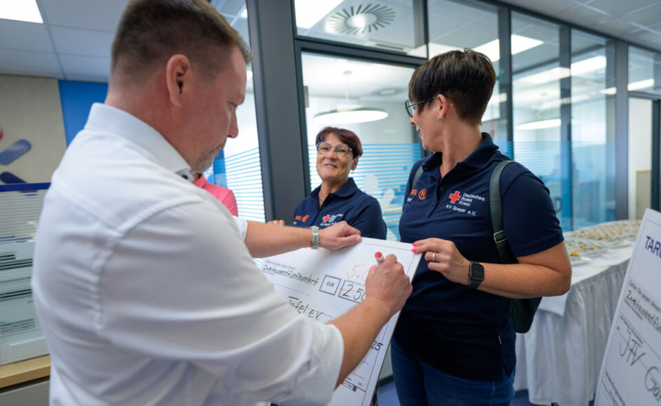 Eine Person in einem weißen Hemd überreicht einen großen Scheck an zwei Frauen in blauen Poloshirts mit dem Logo des Deutschen Roten Kreuzes. Die Frauen lächeln und scheinen sich über den Scheck zu freuen. Im Hintergrund sind weitere Personen und ein Tisch mit Snacks zu sehen.