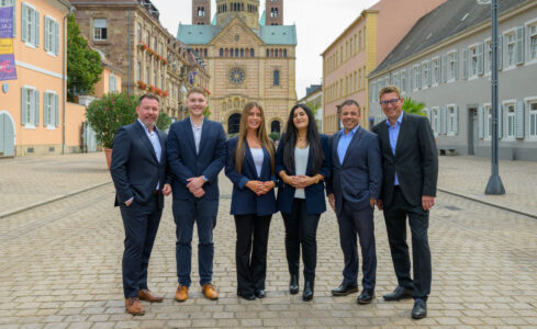 Sechs Personen stehen in formeller Kleidung auf einer gepflasterten Straße, mit einem historischen Gebäude im Hintergrund. Die Gruppe besteht aus drei Männern und drei Frauen. Der Hintergrund zeigt eine charmante Stadtansicht mit bunten Gebäuden und Pflanzen.
