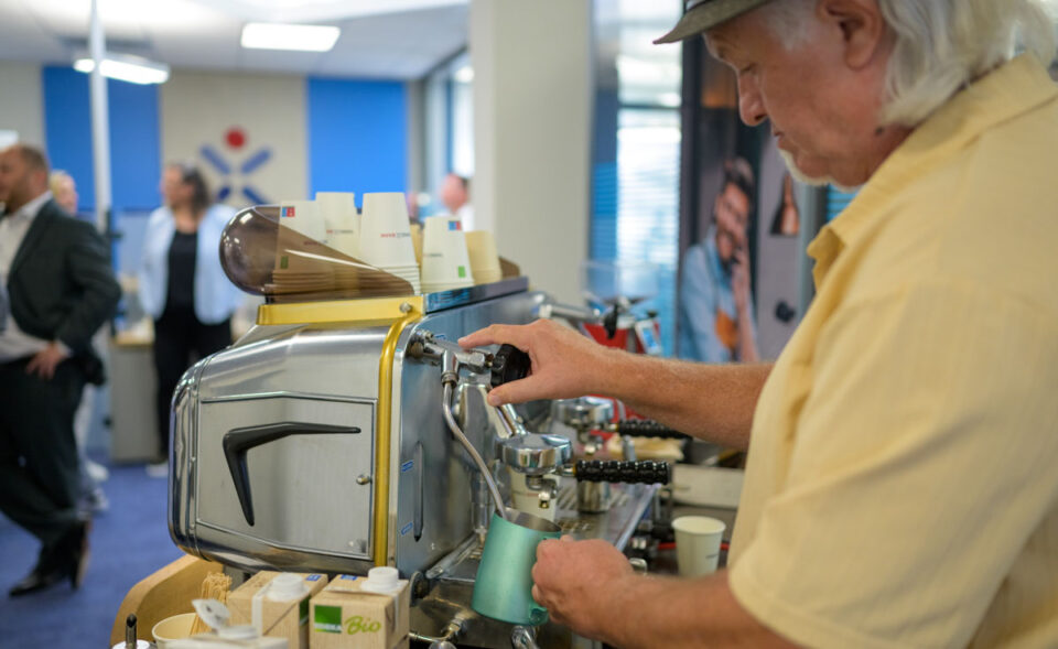 Ein Mann mit langen, weißen Haaren und einem Hut bereitet Kaffee an einer Espressomaschine. Er trägt ein gelbes Hemd und hält eine Kanne, während er die Maschine bedient. Im Hintergrund sind andere Personen und eine moderne Büroumgebung zu sehen.