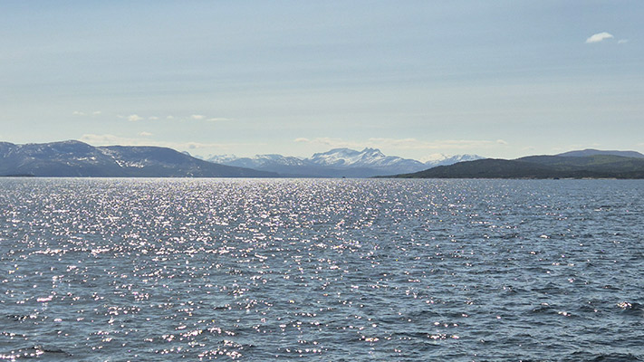 Eine ruhige Wasseroberfläche mit funkelnden Lichtreflexionen. Im Hintergrund sind schneebedeckte Berge und eine klare, hellblaue Himmel zu sehen. Die Landschaft vermittelt eine friedliche Atmosphäre.