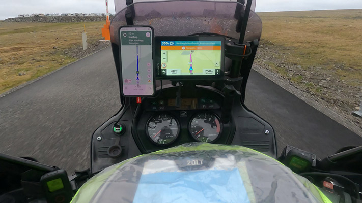Das Bild zeigt die Sicht eines Fahrers auf einem Motorrad. Im Vordergrund sind das Motorrad-Armaturenbrett mit zwei digitalen Displays zu sehen. Links ist ein Smartphone mit Navigationsanweisungen, während das rechte Display Informationen wie Temperatur und Kilometerstand anzeigt. Die Straße führt geradeaus und ist von einer grünen Landschaft umgeben. Es ist bewölkt, und die Straße ist nass.