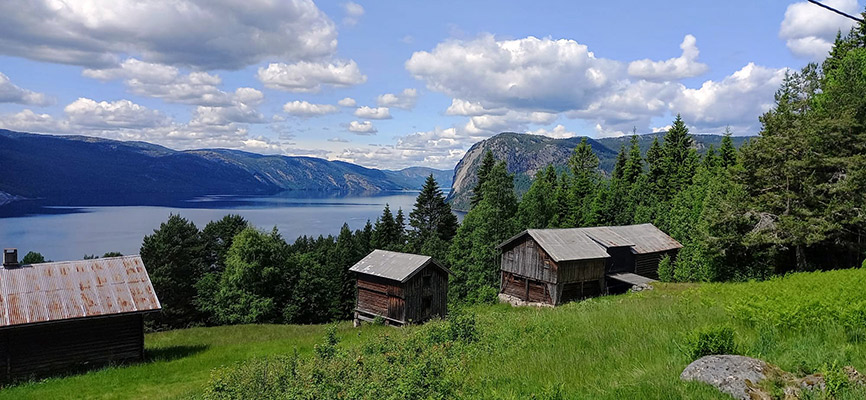 Eine Landschaft mit mehreren alten Holzhäusern im Vordergrund, umgeben von üppigem Grün. Im Hintergrund erstreckt sich ein ruhiger See, umgeben von Bergen und Wäldern unter einem klaren, blauen Himmel mit weißen Wolken.