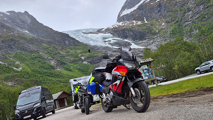 Drei Motorräder stehen auf einer Straße vor einer beeindruckenden Berglandschaft. Im Hintergrund ist ein Gletscher sichtbar, umgeben von grünen Hügeln und steilen Felsen. Das Wetter ist bewölkt und es sieht kühl aus. Ein großer, dunkler Lieferwagen parkt in der Nähe, sowie ein weiteres Fahrzeug weiter rechts. Auf der linken Seite ist ein Informationsschild zu sehen.