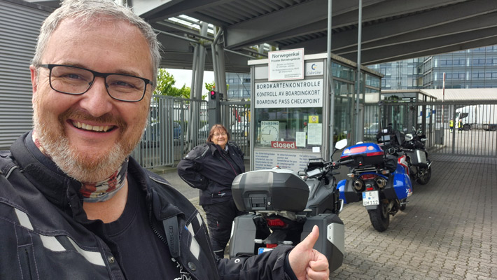 Ein Mann mit Brille und Bart lächelt in die Kamera und zeigt mit dem Daumen nach oben. Im Hintergrund steht eine Frau, die ebenfalls lächelt. Sie befinden sich an einem Kontrollpunkt mit einem Schild, das 