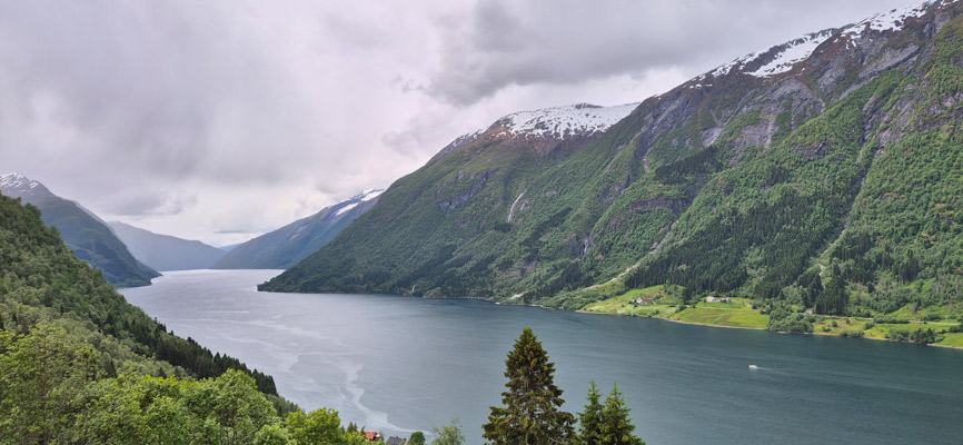 Eine panoramische Ansicht eines Fjords umgeben von hohen, bewaldeten Bergen mit schneebedeckten Gipfeln. Der Himmel ist bewölkt und das Wasser ist ruhig, mit einigen kleinen Booten auf der Oberfläche. In der Ferne sind grüne Wiesen und kleine Häuser sichtbar.