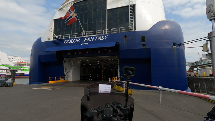 Das Bild zeigt das Fährschiff COLOR FANTASY im Hafen. Die große, blaue Schiffsseite hat große Fenster und ein offenes Garagentor. Im Hintergrund sind andere Schiffe sichtbar. Die Atmosphäre ist hell und klar, mit einem leicht bewölkten Himmel.