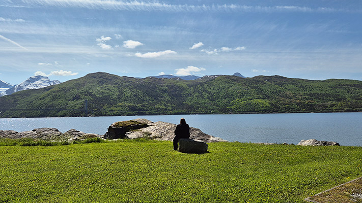 Eine Person sitzt auf einem Felsen am Ufer eines ruhigen Wassers, umgeben von grünen Bergen und blauen Himmel. Die Landschaft ist friedlich.