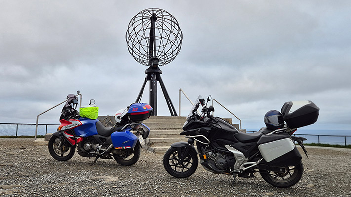 Zwei Motorräder stehen vor einem großen globusförmigen Monument auf einer Plattform. Das eine Motorrad ist rot und blau mit einem grünen Gepäckstück, das andere ist schwarz mit einem grauen Koffer. Der Himmel ist bewölkt, und im Hintergrund ist das Meer sichtbar.