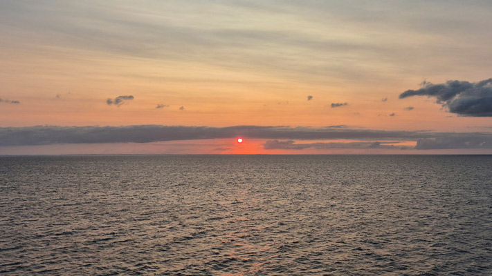 Ein Sonnenuntergang über dem Meer mit sanften Wellen. Die Sonne ist rot und geht hinter einer Wolkenbank unter, während der Himmel in sanften Orange- und Blautönen leuchtet.