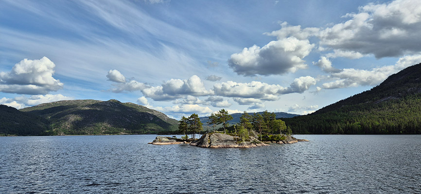 Eine ruhige Landschaft mit einem kleinen, bewaldeten Inselchen in der Mitte eines großen Gewässers. Im Hintergrund sind sanfte Hügel und Berge zu sehen, unter einem Himmel mit vielen Wolken. Die Wasseroberfläche reflektiert die umgebende Natur.