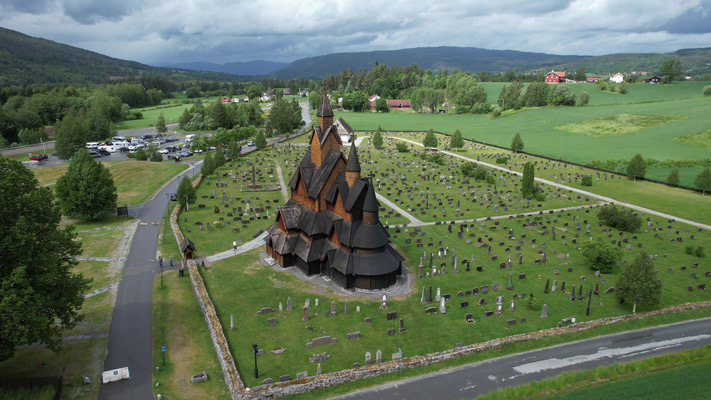 Eine beeindruckende Holzkirche im Stabkirchenstil, umgeben von einem weitläufigen Friedhof mit vielen Grabsteinen. Im Hintergrund erstrecken sich grüne Wiesen und Hügel, während sich am Horizont Berge erheben. Der Himmel ist bewölkt und es sind einige Autos auf einem Parkplatz sichtbar.