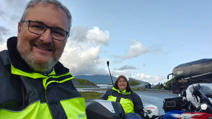 Zwei Personen in leuchtend gelben Motorradjacken posieren für ein Selfie. Im Hintergrund sind Motorräder und eine Busfahrzeug sichtbar. Die Szene zeigt eine kurvenreiche Straße und Wolken am Himmel.