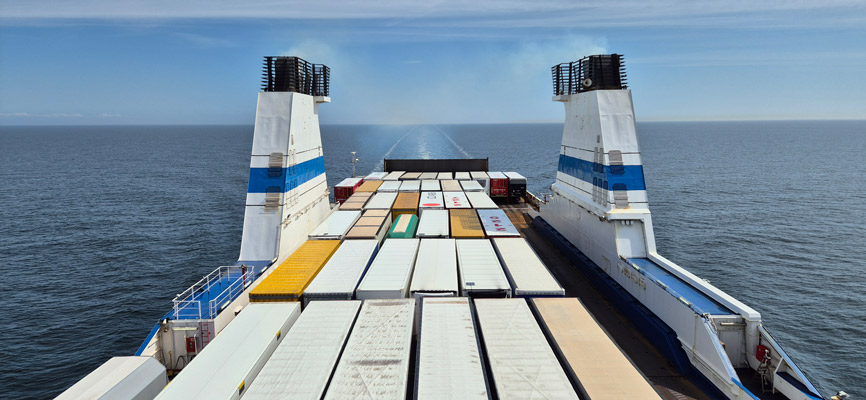 Ein Blick von der Oberseite eines Frachtschiffs auf das offene Meer. Die Decks sind mit verschiedenen Containern in unterschiedlichen Farben und Größen beladen. Im Hintergrund sind die Wellen des Wassers zu sehen, während das Schiff sich fortbewegt. Der Himmel ist klar mit wenigen Wolken.