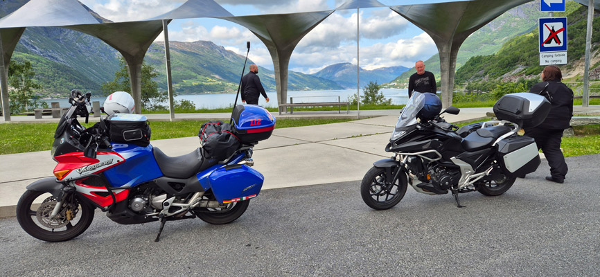 Zwei Motorräder stehen auf einem Parkplatz mit Blick auf einen Fjord. Das linke Motorrad ist rot und blau, das rechte schwarz und grau. Im Hintergrund sind Berge und ein See sichtbar. Zwei Personen gehen in Richtung Wasser, während eine weitere Person neben dem Motorrad steht.