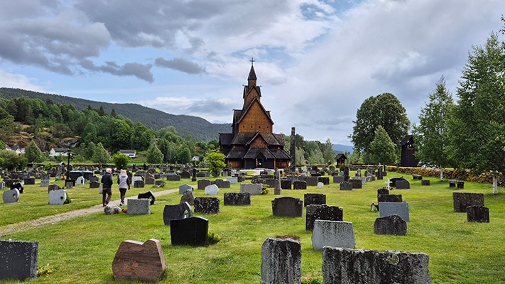 Eine weitläufige Friedhofslandschaft mit vielen Grabsteinen in verschiedenen Formen und Größen. Im Hintergrund erhebt sich eine beeindruckende Holzkirche mit einem spitzen Turm, umgeben von Bäumen und Hügeln. Der Himmel ist bewölkt, und einige Spaziergänger sind auf dem Weg durch den Friedhof zu sehen.