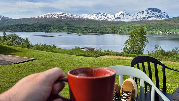 Eine Person hält eine rote Tasse in der Hand, während sie auf einer Terrasse sitzt. Im Hintergrund erstreckt sich ein ruhiger See mit schneebedeckten Bergen und grünen Hügeln. Die Landschaft ist üppig und friedlich, mit Bäumen auf der rechten Seite und einem klaren Himmel. Die Füße der Person sind in braunen Schuhen, die auf einem Stuhl ruhen.