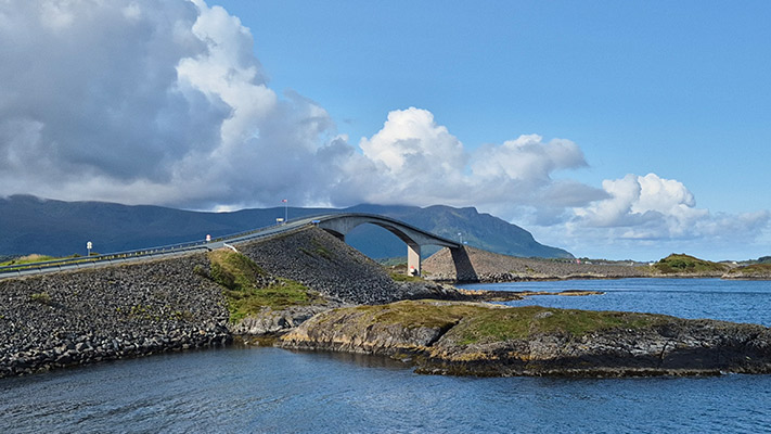 Ein Bogenbrücke, die eine kleine Insel mit dem Festland verbindet, umgeben von Wasser und Bergen unter einem blauen Himmel mit Wolken.