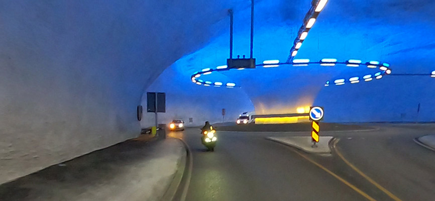 Ein Tunnel mit blauer Beleuchtung, in dem ein Motorrad und einige Autos fahren. Der Tunnel hat eine runde Form mit einer Leuchte an der Decke und Verkehrsschildern an den Wänden. Die Straße führt in eine Kurve.