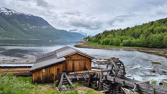 Eine Holzstruktur mit einem Wasserrad steht am Ufer eines ruhigen Gewässers. Im Hintergrund sind grüne Hügel und schneebedeckte Berge sichtbar, unter einem bewölkten Himmel. Die Landschaft ist üppig und naturnah.