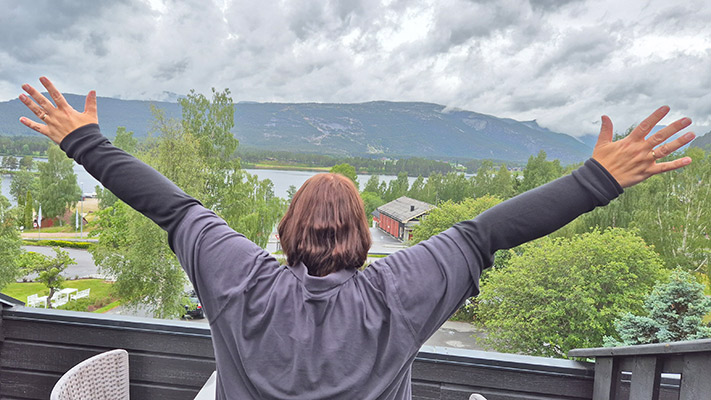 Eine Person steht mit ausgebreiteten Armen auf einem Balkon und schaut auf eine Landschaft mit Bergen, Bäumen und einem Gewässer. Der Himmel ist bewölkt.