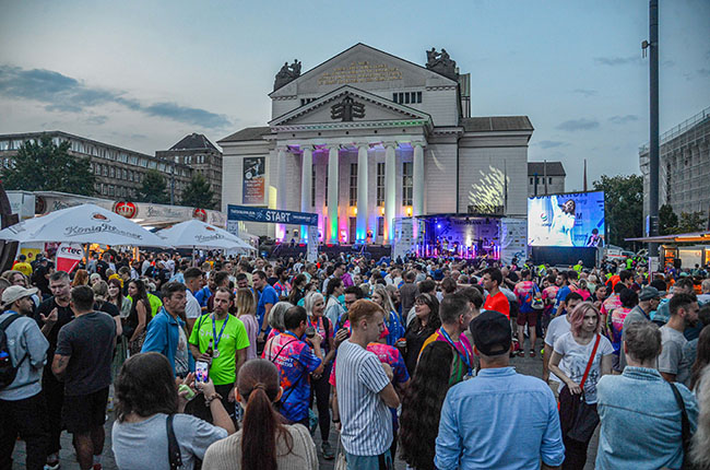 Eine große Menschenmenge versammelt sich vor einem beeindruckenden Gebäude mit Säulen. Es gibt eine Bühne mit Lichtern und Bildschirmen, die Musik spielen. Die Anwesenden tragen bunte Kleidung und scheinen an einem Festival oder Event teilzunehmen. Im Hintergrund sind weitere Menschen und Verkaufsstände sichtbar.