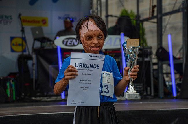 Ein junger Teilnehmer hält stolz eine Urkunde mit dem Namen Abdallah Yassir und einen Pokal in der Hand. Er trägt ein blaues T-Shirt und steht auf einer Bühne mit einem DJ im Hintergrund. Die Stimmung ist festlich, und im Hintergrund sind Lichter zu sehen.