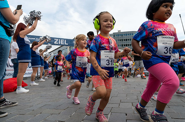 Eine Gruppe von Kindern läuft fröhlich in einem Rennen. Einige tragen bunte T-Shirts mit dem Logo von TARGOBANK. Die Kinder haben Nummern auf ihren Shirts und eine der Mädchen trägt Kopfhörer. Im Hintergrund jubeln Menschen und es gibt ein Banner mit der Aufschrift 'ZIEL'.