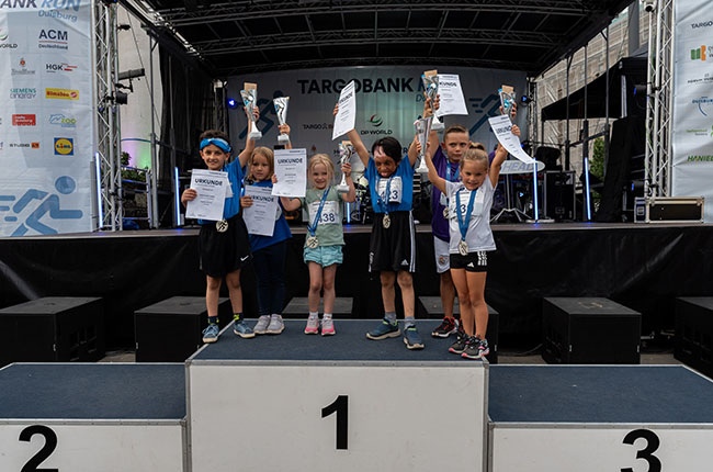 Eine Gruppe von sieben Kindern steht auf einem Siegertreppchen, jeder hält eine Urkunde und eine Medaille. Die Kinder tragen sportliche Kleidung, darunter T-Shirts und Shorts. Im Hintergrund sind Banner mit Sponsorenlogos sichtbar. Die Kinder scheinen glücklich und stolz über ihre Auszeichnungen zu sein. Die Szene findet während einer Sportveranstaltung statt.