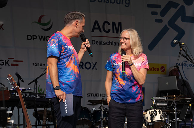 Zwei Personen stehen auf einer Bühne und sprechen miteinander. Beide tragen t-shirts in bunten Farben mit blau-orange-rotem Muster. Der Mann hält ein Mikrofon in der linken Hand und schaut die Frau an, die ebenfalls ein Mikrofon in der rechten Hand hält. Im Hintergrund sind Musikinstrumente und eine große Leinwand mit Logos sichtbar.