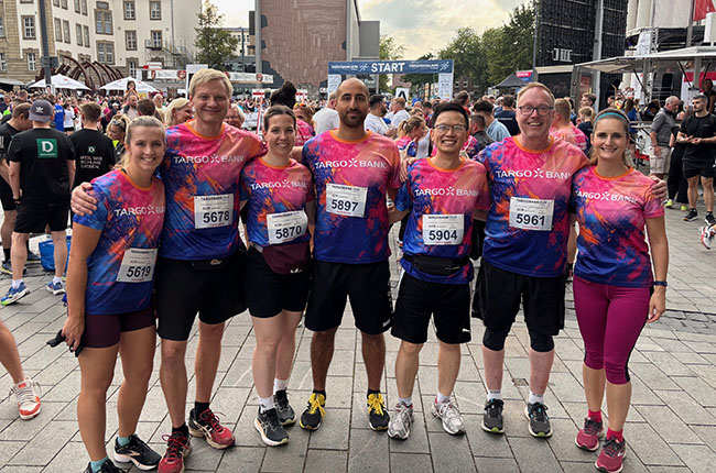 Eine Gruppe von sieben Personen steht zusammen, gekleidet in bunte T-Shirts mit dem Aufdruck 