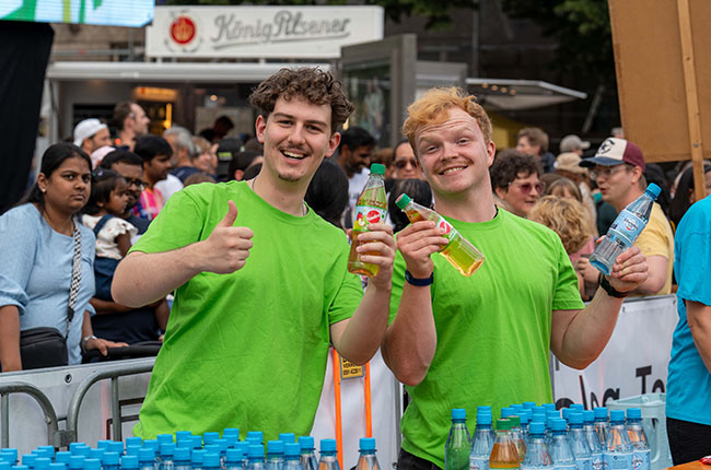 Zwei junge Männer in grünen T-Shirts stehen vor einer Menschenmenge. Einer zeigt den Daumen hoch und hält eine Flasche in der Hand, während der andere ebenfalls eine Flasche hält und lächelt. Im Hintergrund sind Menschen und ein Verkaufsstand sichtbar.