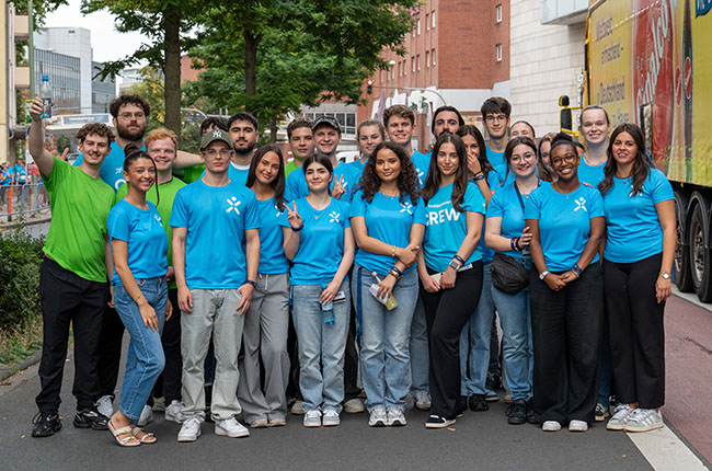 Eine Gruppe von jungen Menschen steht in blauen T-Shirts mit der Aufschrift „CREW“ zusammen. Sie posieren für ein Foto auf einer Straße, umgeben von Bäumen und Gebäuden. Einige Mitglieder halten Getränke in der Hand, während andere lächeln oder Peace-Zeichen zeigen. Die Gruppe hat eine freundliche und einladende Ausstrahlung.