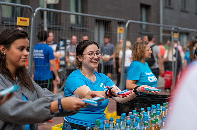 Zwei Frauen in blauen T-Shirts stehen an einem Tisch mit Getränken und Snacks. Eine der Frauen, mit Brille, lächelt und reicht einen Snack. Im Hintergrund sind viele Menschen zu sehen, die auf ein Event warten.
