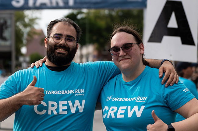 Zwei Personen stehen lächelnd vor einem Startbanner. Sie tragen T-Shirts mit der Aufschrift 'TARGOBANK RUN Duisburg CREW' und zeigen Daumen hoch.